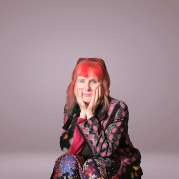 Michelle Baharier surrounded by her artwork (canvases). She has dyslexia. Michelle is wearing lots of colours and has dyed red hair. She is smiling shyly.